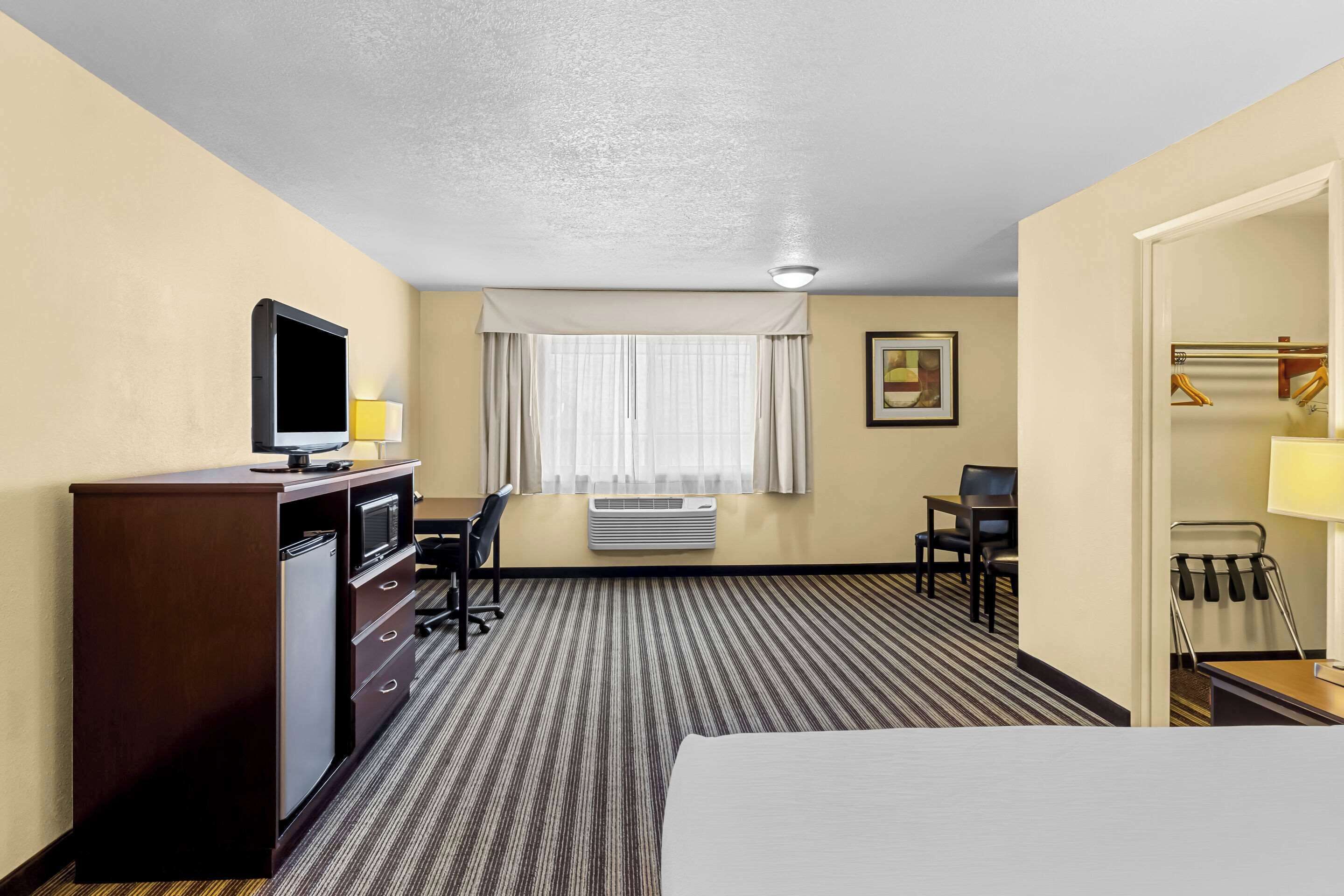 A hotel room with a bed, a desk with a computer, a TV, a minibar, a coat rack, and a window with curtains.