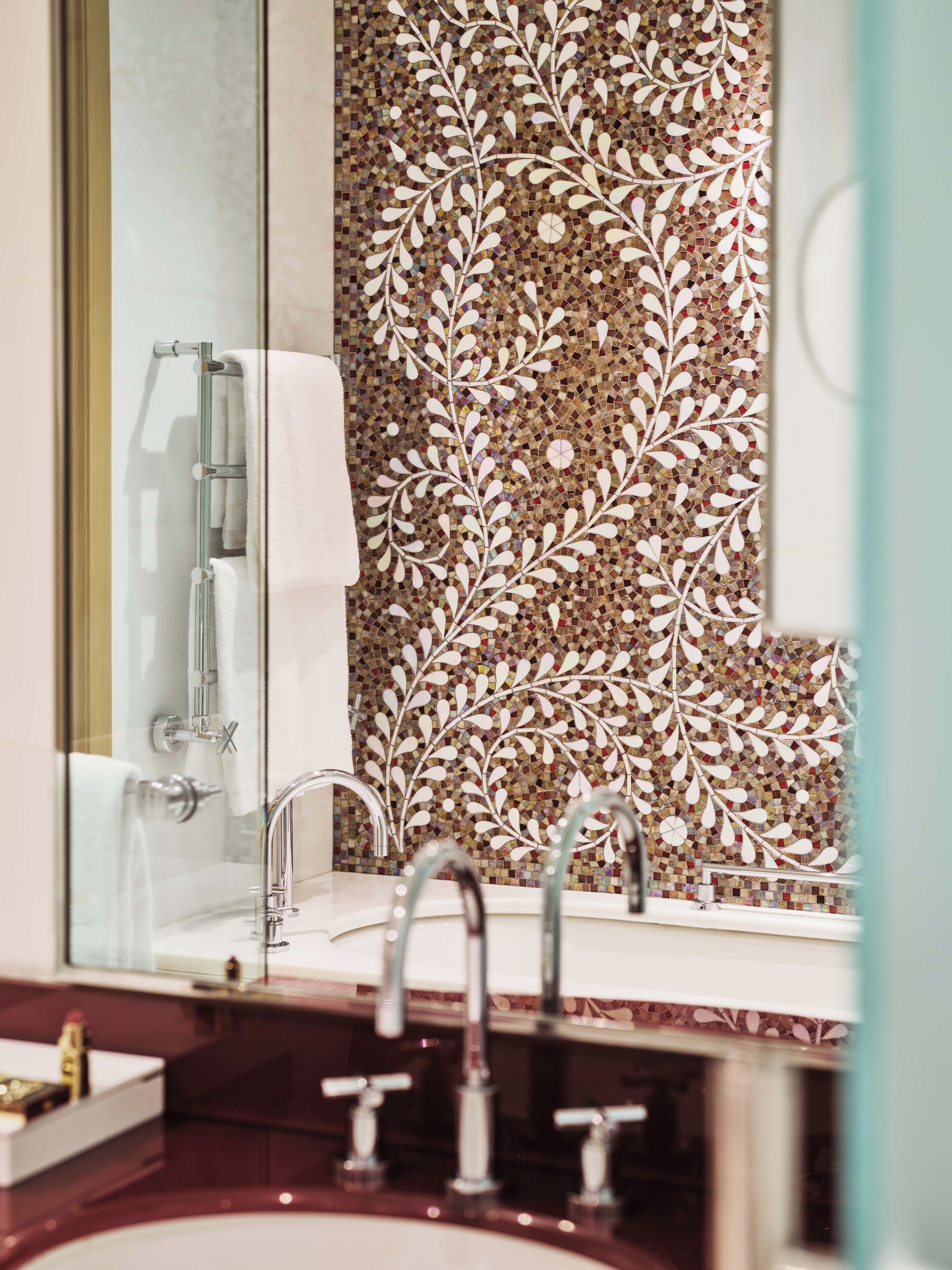 A bathroom with a glass shower enclosure, a white towel, a sink with two faucets, and a decorative mosaic wall.