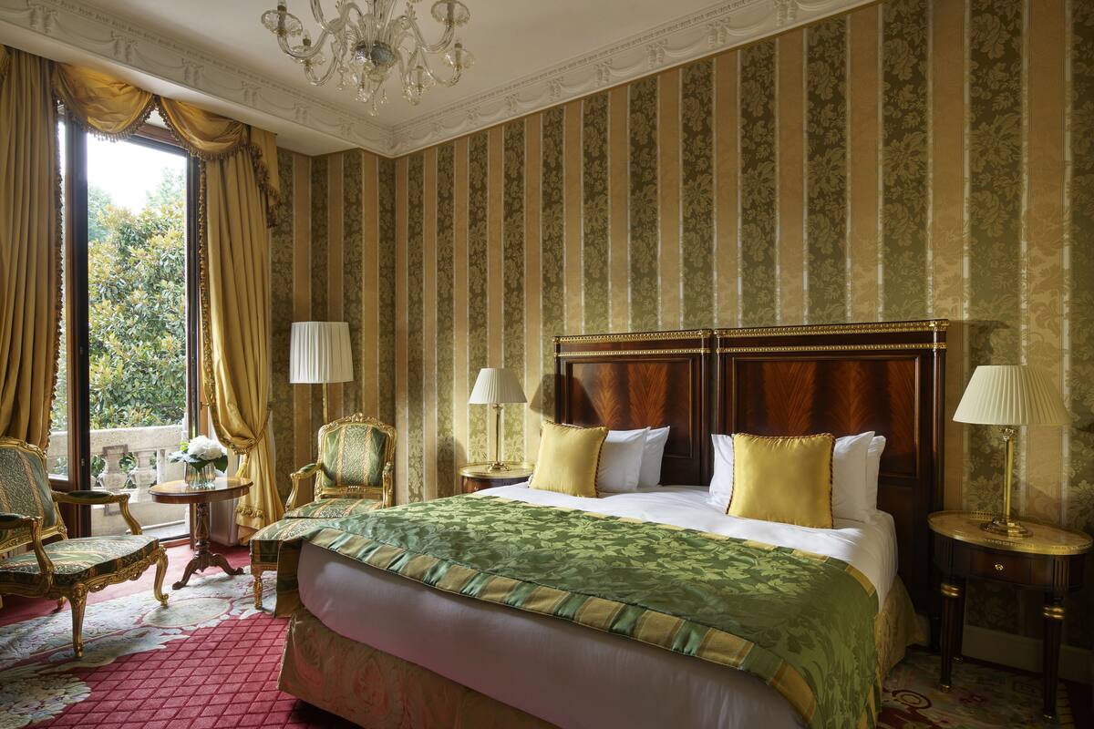 A spacious bedroom with a large bed, ornate headboard, and a view of greenery outside.