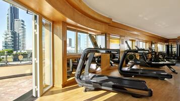 Ein Fitnessstudio mit Laufbändern, Trainingsgeräten und großen Fenstern mit Blick auf die Stadt.