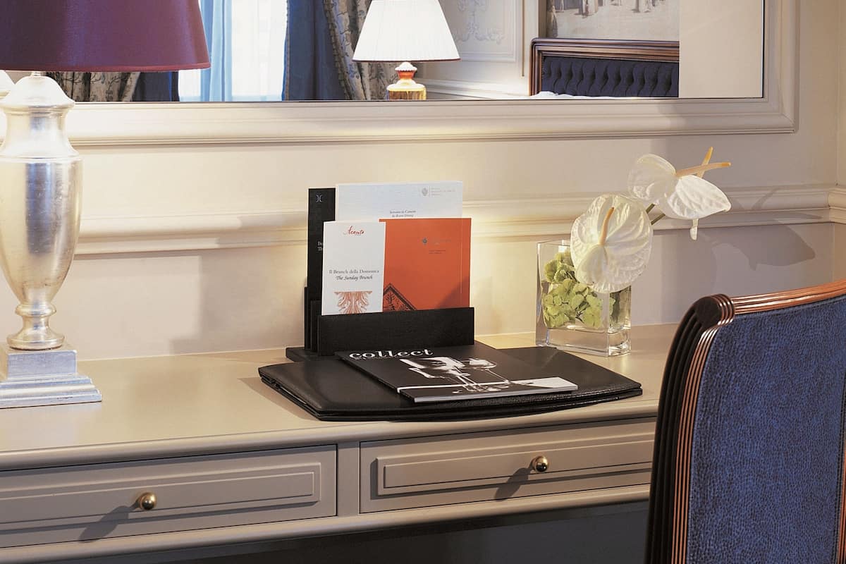 A hotel room with a desk, a lamp, a framed picture, and a vase with flowers.