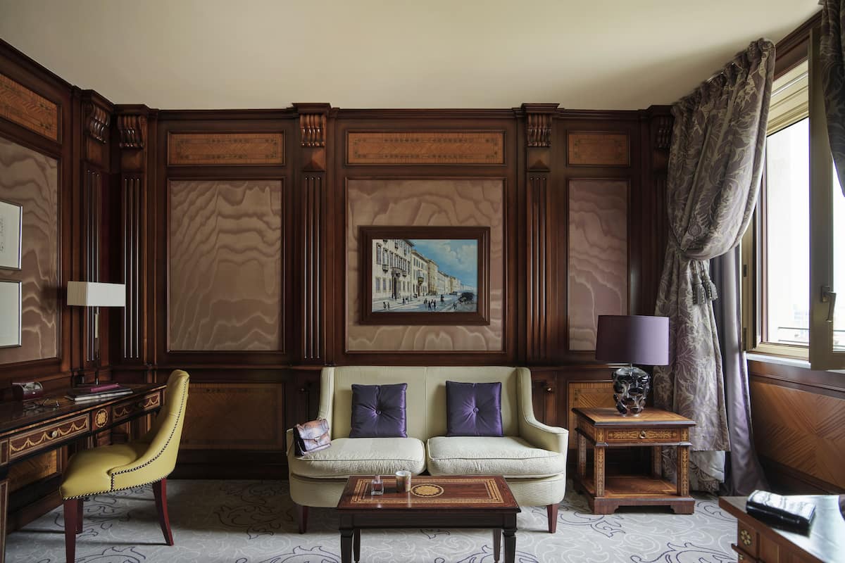 A room with a sofa, armchair, and a desk with a lamp. There is a painting on the wall and a window with curtains.