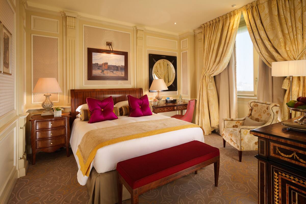 A hotel room with a large bed, a wooden nightstand, a desk, a chair, and a window with curtains.