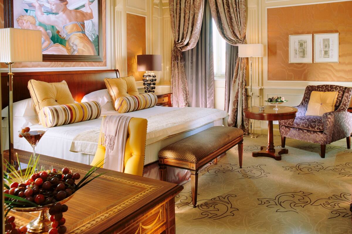 A luxurious hotel room with a large bed, a sitting area with a chair and table, a painting on the wall, and a window with curtains.