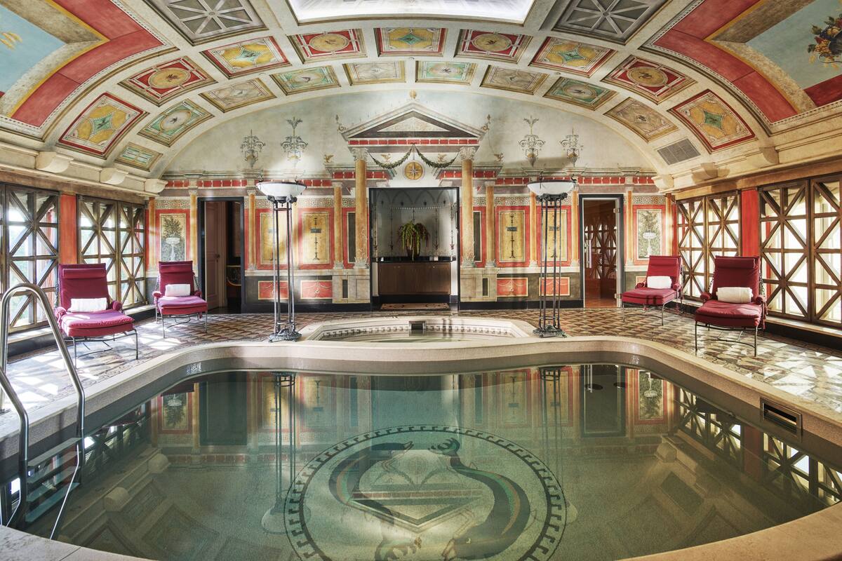 A luxurious indoor pool area with a central fountain, red and gold decorative elements, and a high ceiling with skylights.