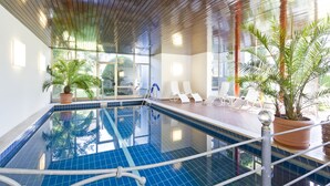 An indoor swimming pool with clear blue tiles, surrounded by lounge chairs and potted plants.