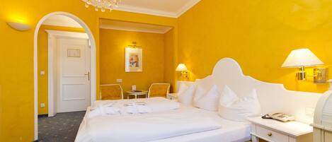 Habitación de hotel con una cama grande, un escritorio y una silla. Se ve una puerta que conduce a otra habitación y un cuadro en la pared.