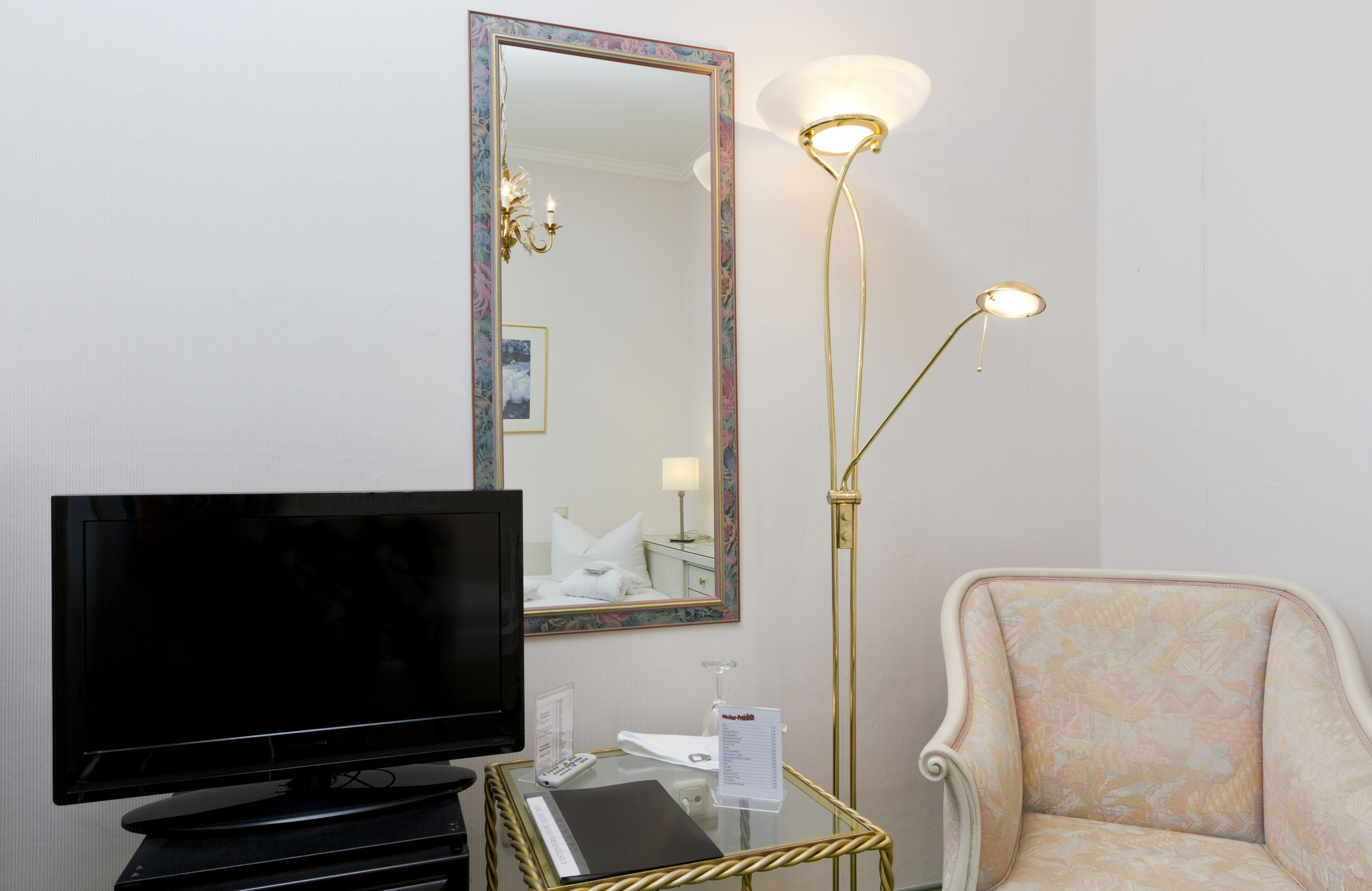 Habitación de hotel con televisor de pantalla plana, espejo con marco dorado, lámpara de pie y sillón con estampado.