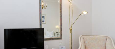 Habitación de hotel con televisor de pantalla plana, espejo con marco dorado, lámpara de pie y sillón con estampado.