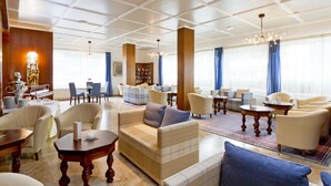 A spacious hotel lobby with a high ceiling, a central seating area with sofas and armchairs, a dining area with a table and chairs, and a large bookshelf.