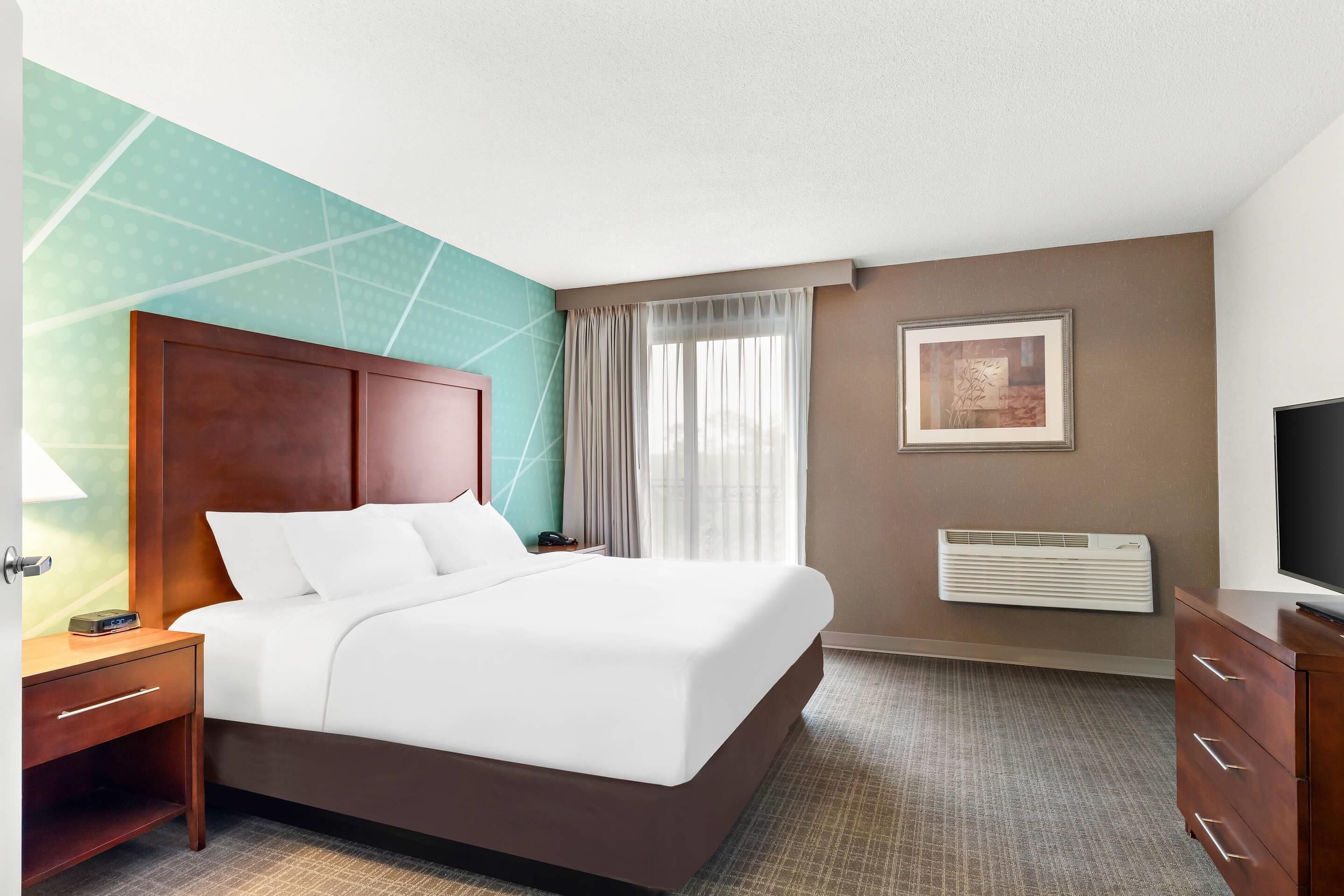A hotel room with a bed, bedside table, lamp, air conditioning unit, and a television.