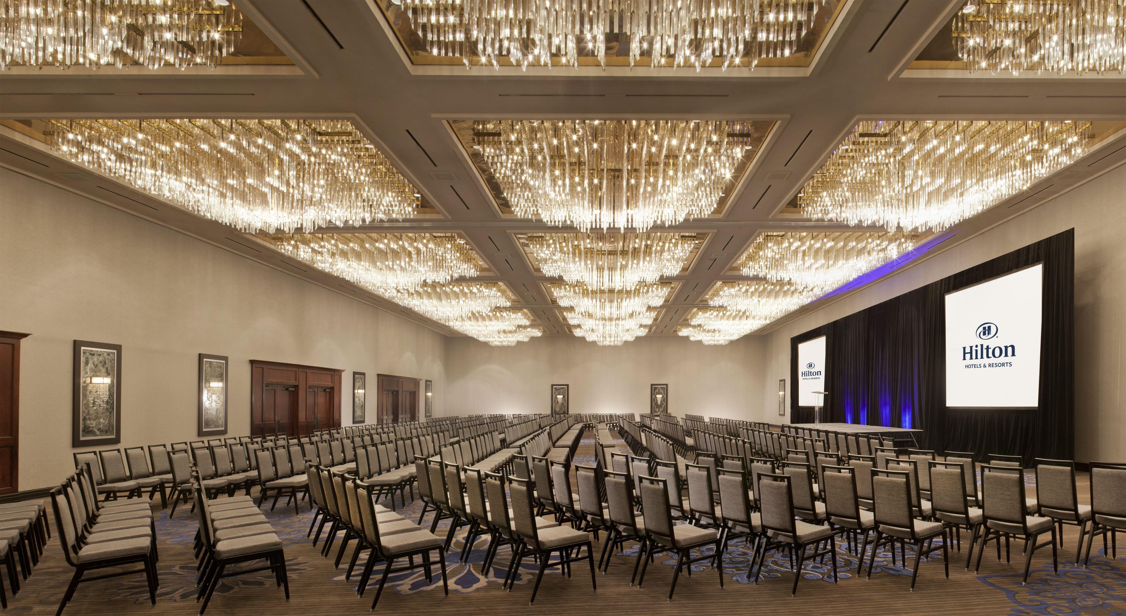 Un salón de conferencias con filas de sillas, una pantalla grande mostrando el logotipo de Hilton y una araña con múltiples lámparas.
