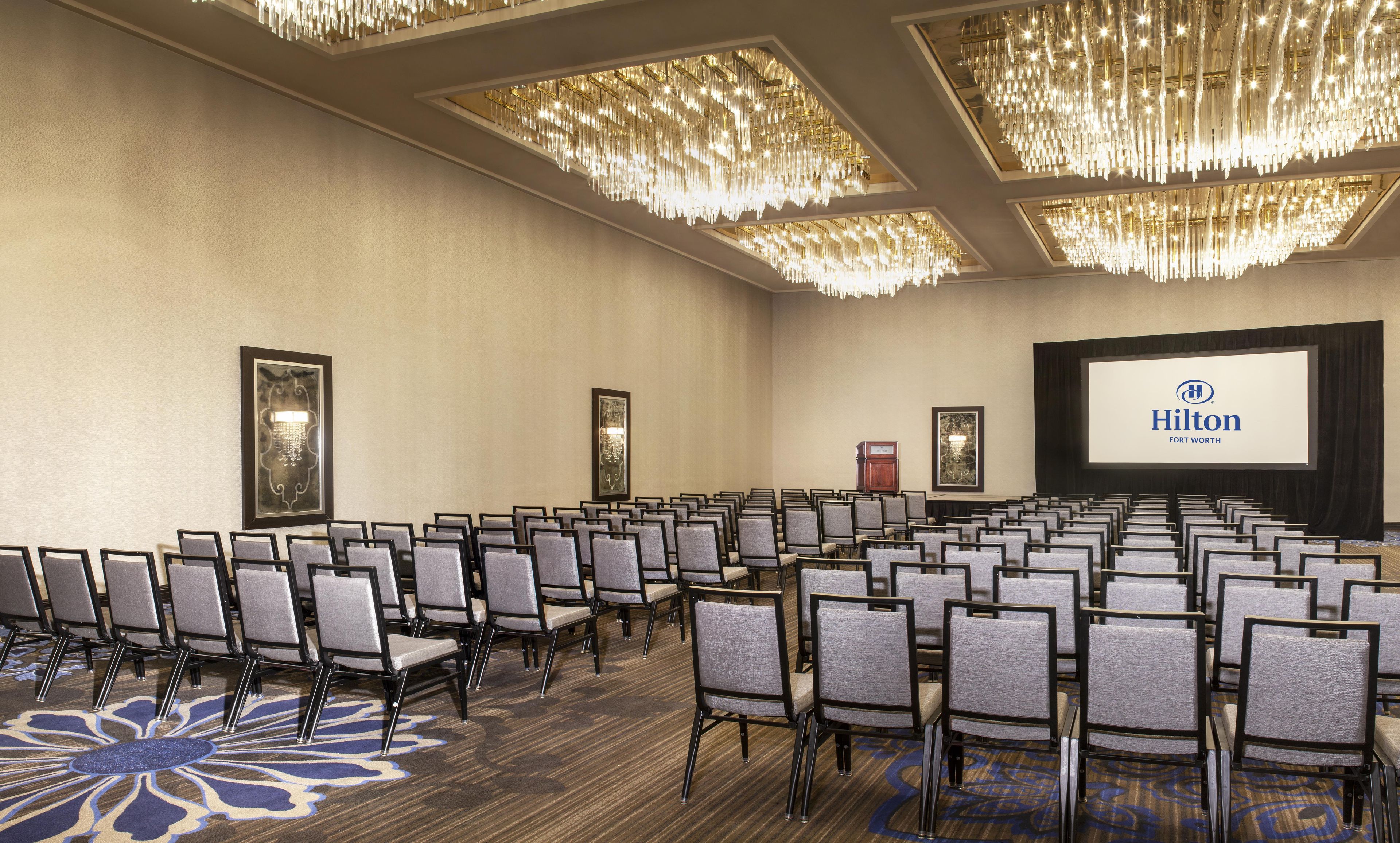 Un salón de conferencias con filas de sillas, una pantalla grande mostrando el logotipo de Hilton y candelabros ornamentales.