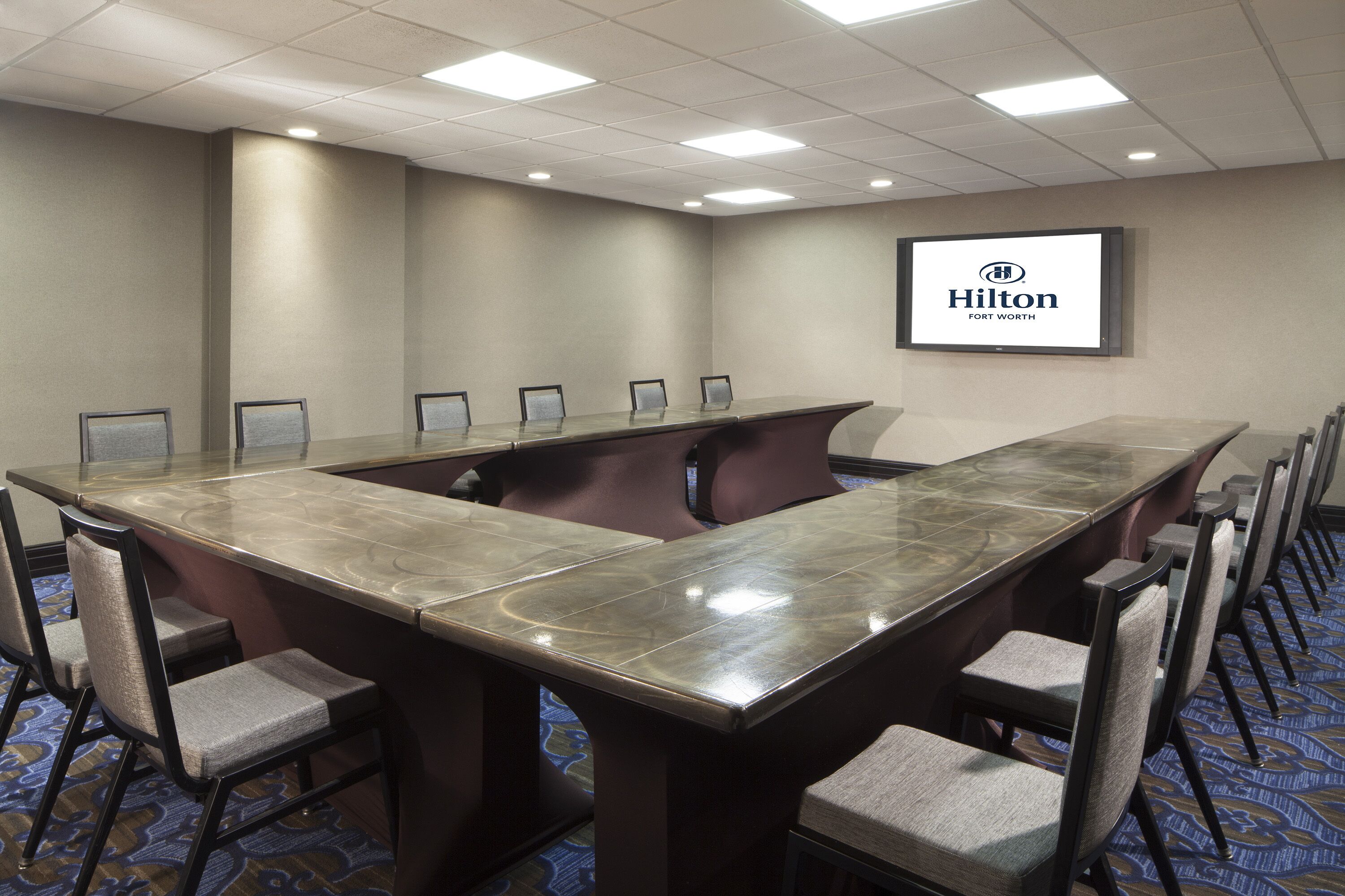 A conference room with a long table, chairs, and a screen displaying 'Hilton Fort Worth'.