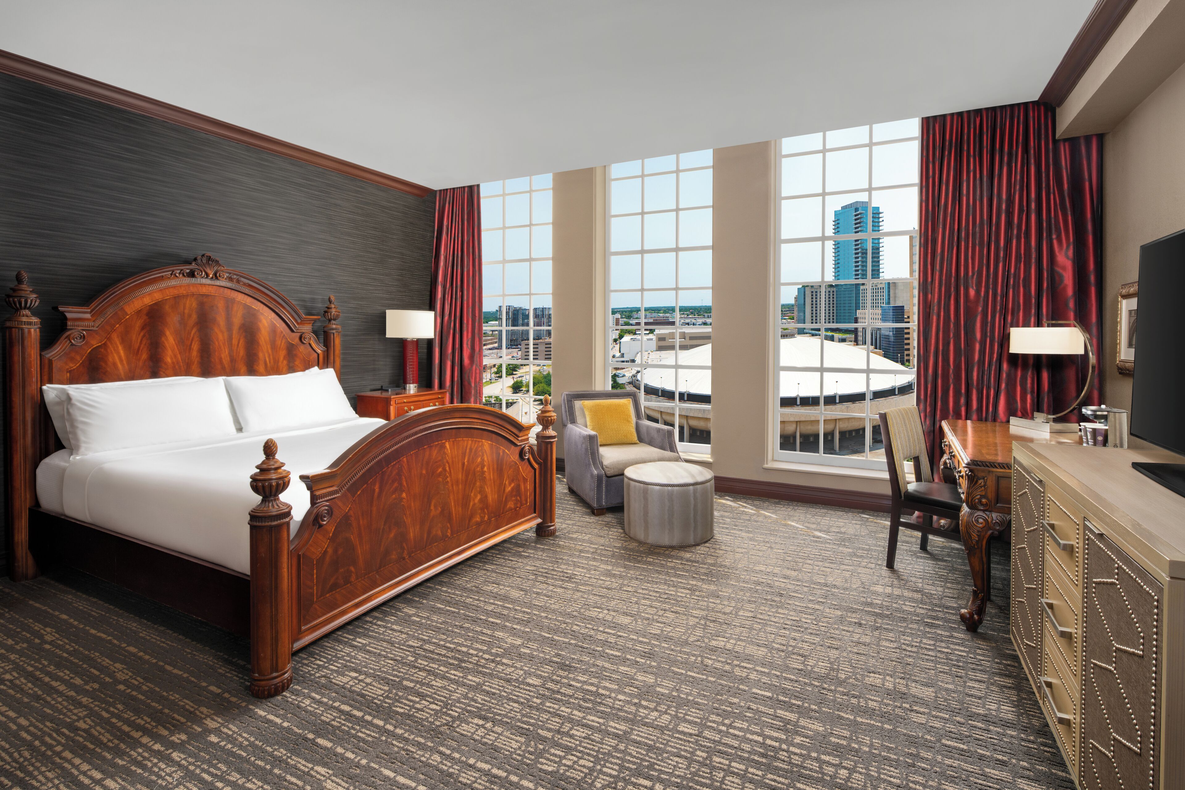 Habitación de hotel con una cama grande, un escritorio, una silla, un otomano pequeño y redondo, y vistas de la ciudad a través de ventanas amplias.