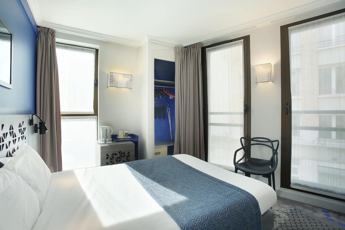 Quarto de hotel com uma cama grande, uma escrivaninha com luminária, uma cadeira e um guarda-roupa.
