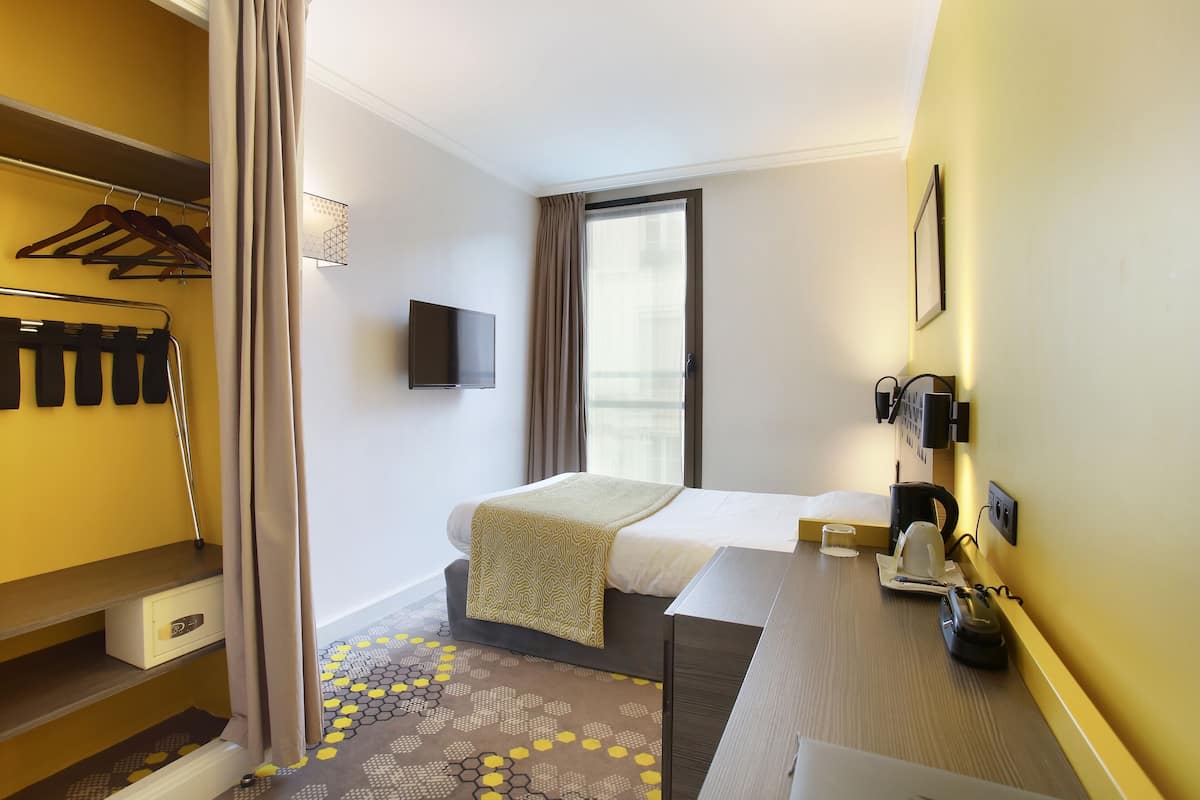 Quarto de hotel com cama, escrivaninha, cadeira e televisão na parede.