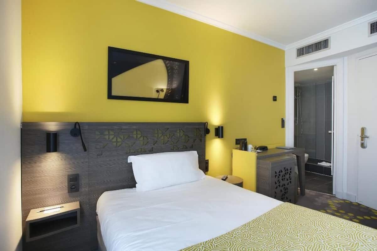 Quarto de hotel com cama, mesa de cabeceira, luminária e espelho na parede.