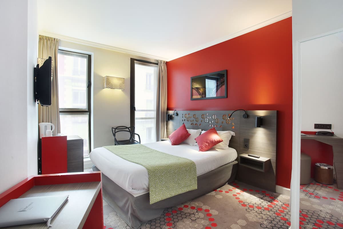 Quarto de hotel com cama, escrivaninha, cadeira, televisão e janela com cortinas.