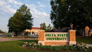 Un cartel de la Universidad Estatal Delta, con base de ladrillo y letras verdes, rodeado de un césped cuidado y plantas con flores.