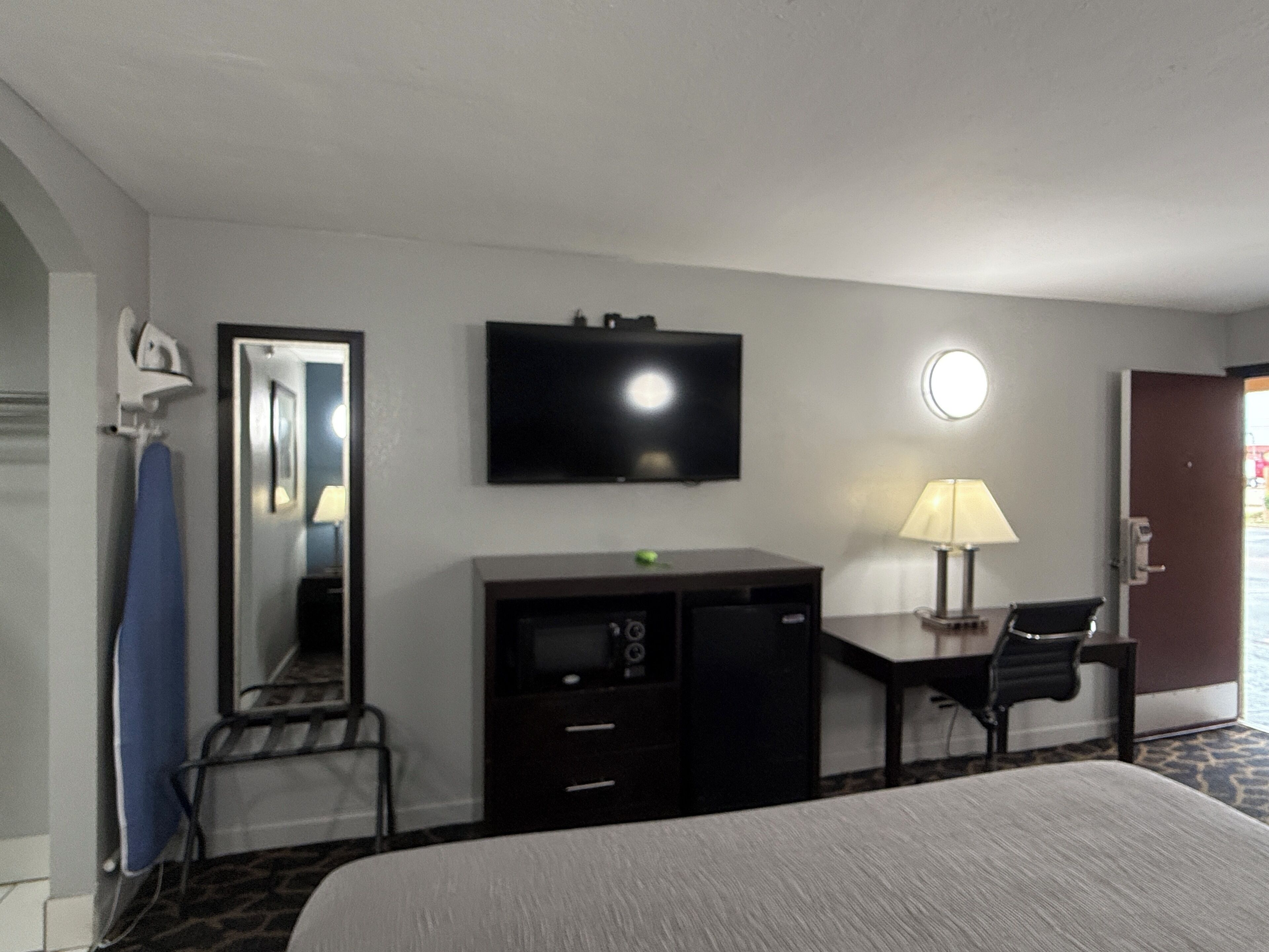 Quarto de hotel com cama, escrivaninha, televisão, abajur e espelho.