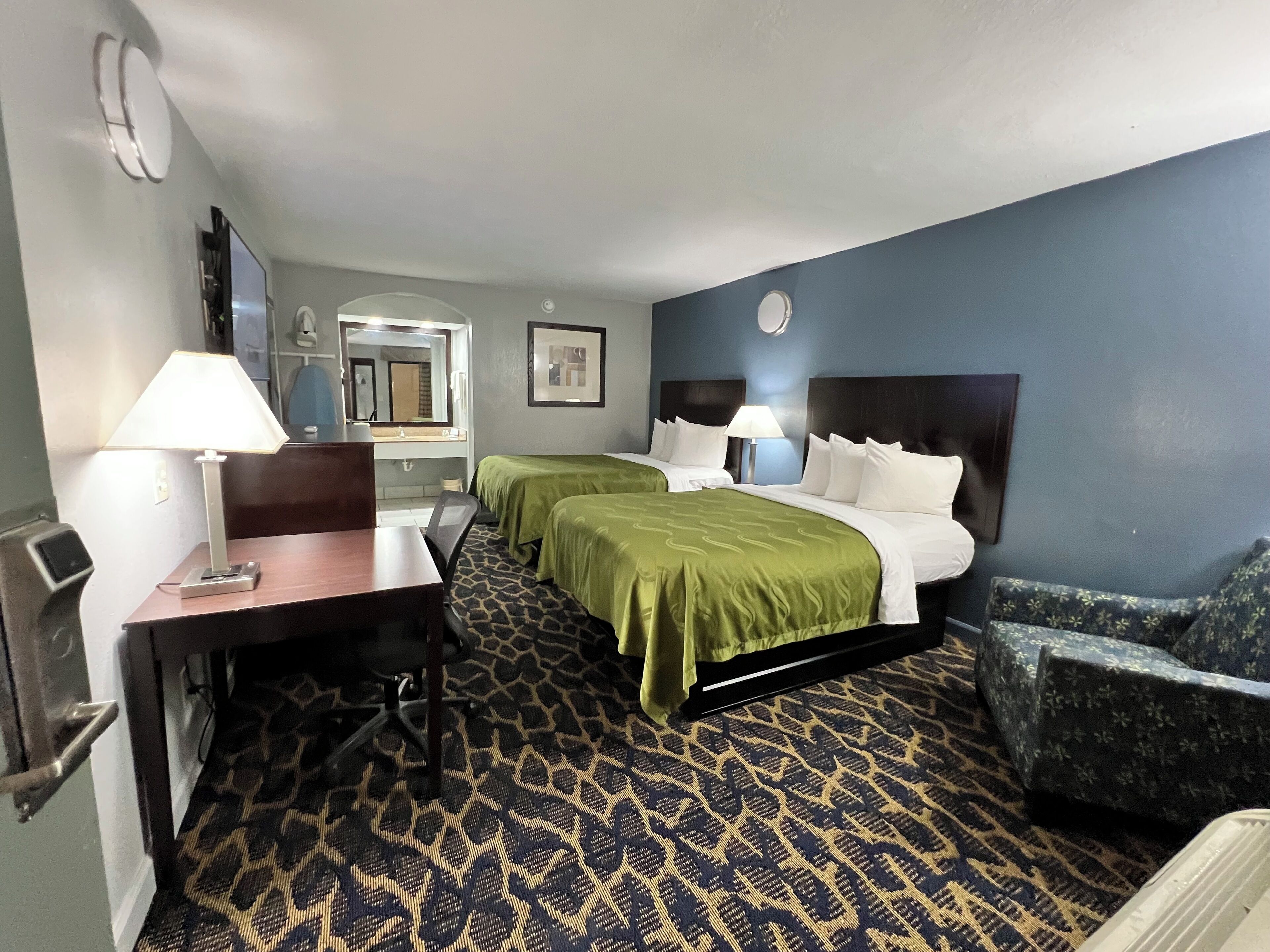 Habitación de hotel con dos camas individuales, un escritorio con silla, una mesita pequeña, un sofá y una alfombra estampada.