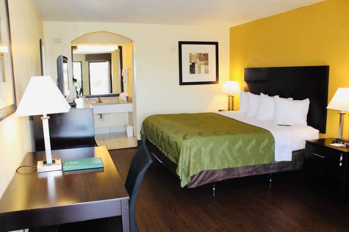 Een hotelkamer met een bed, nachtkastjes, een bureau, een stoel, een spiegel en een lijst met een foto aan de muur.