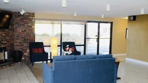 Una sala de estar con un sofá azul, una chimenea, un televisor montado en una pared de ladrillo y un ventanal con vistas al exterior.