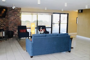Una sala de estar con un sofá azul, una chimenea, un televisor montado en una pared de ladrillo y un ventanal con vistas al exterior.