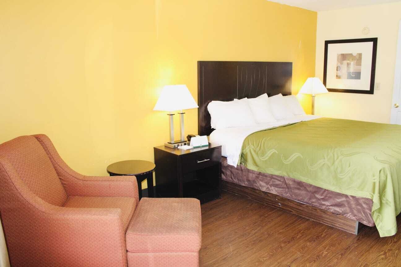 A hotel room with a bed, a chair, a nightstand with a lamp, and a framed picture on the wall.