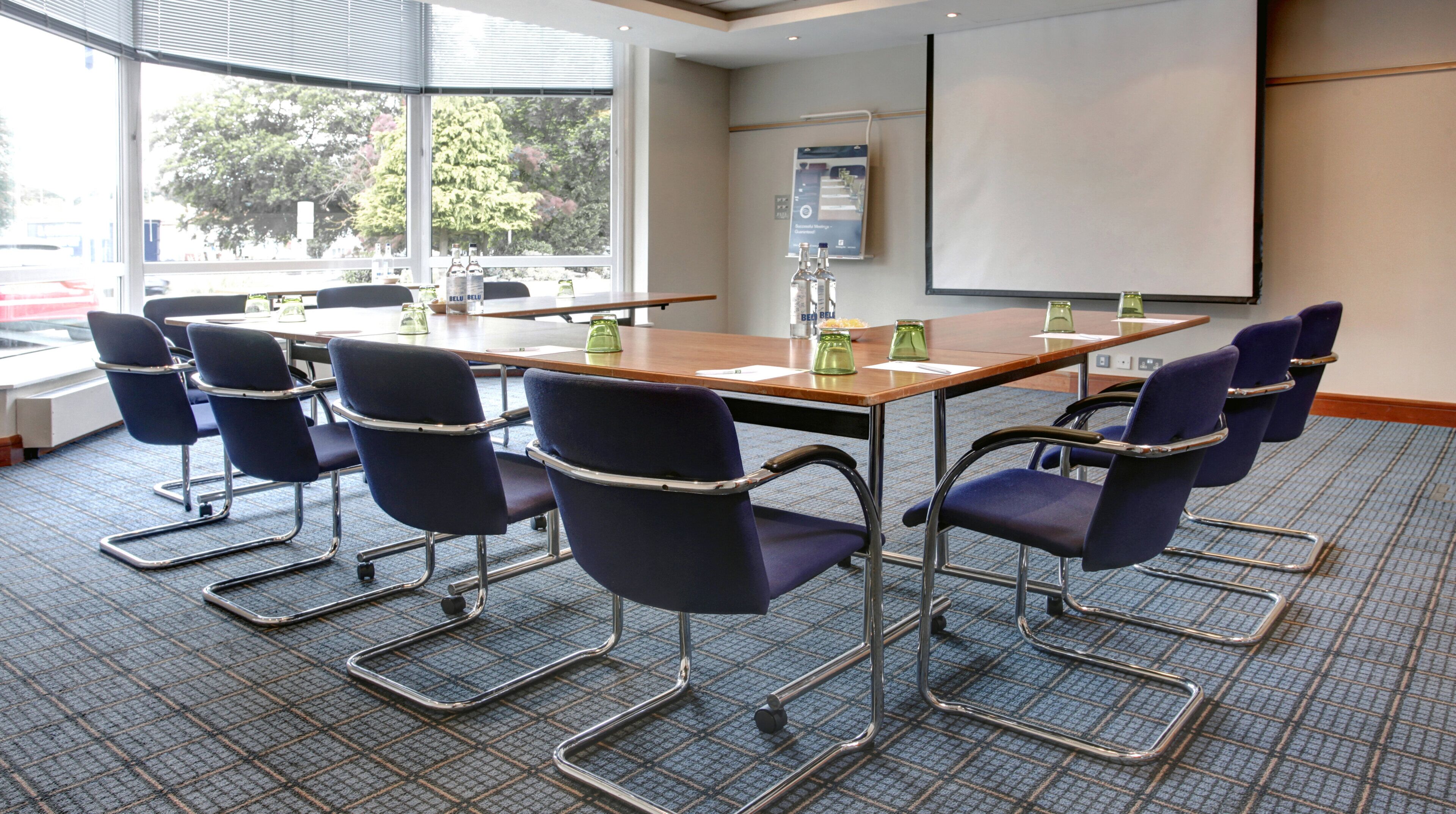 A conference room with a large table, six chairs, a projector screen, and a window with a view of trees.