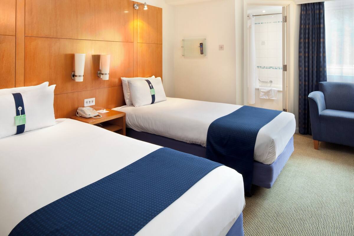 A hotel room with two beds, a wooden headboard, bedside lamps, a desk, a chair, and a bathroom visible through an open door.