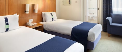 A hotel room with two beds, a wooden headboard, a desk with a phone, a chair, and a window with curtains.