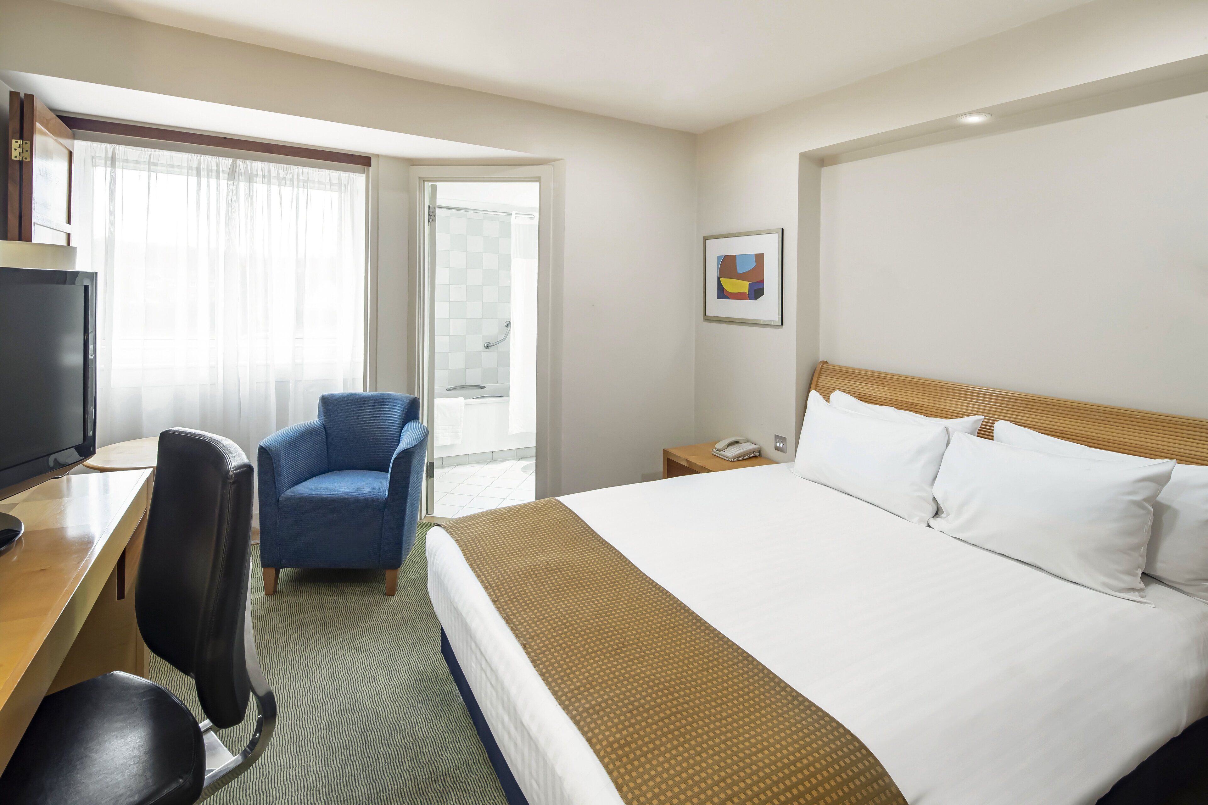 A hotel room with a bed, a desk with a computer, a chair, a phone, and a window with curtains.