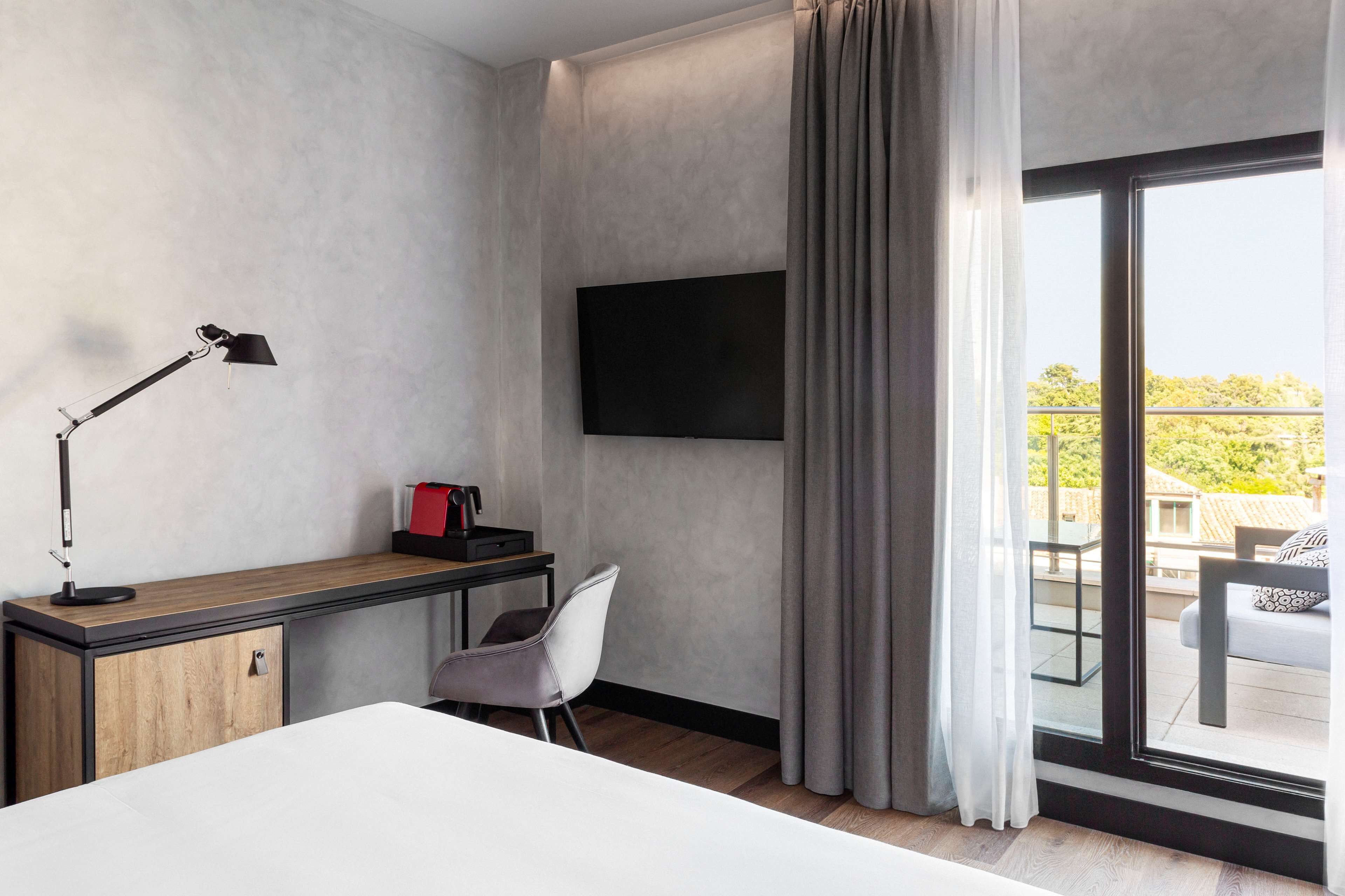 Ein modernes Hotelzimmer mit einem Bett, einem Schreibtisch mit Lampe, einem Sessel, einem Flachbildfernseher und einem Balkon mit Sitzgelegenheit.