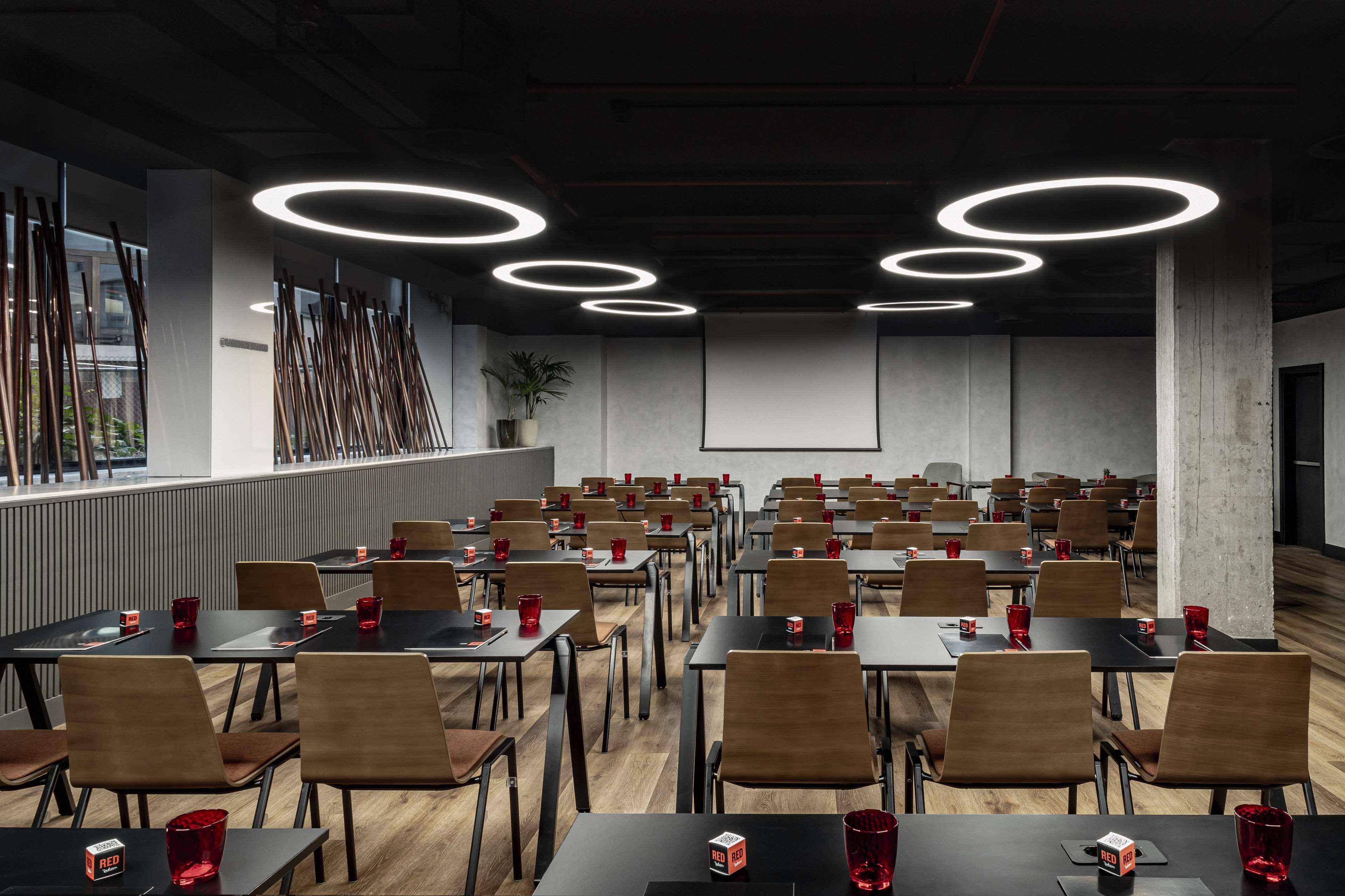 Una sala conferenze con diversi tavoli e sedie, luci a soffitto circolari e un grande schermo di fronte.