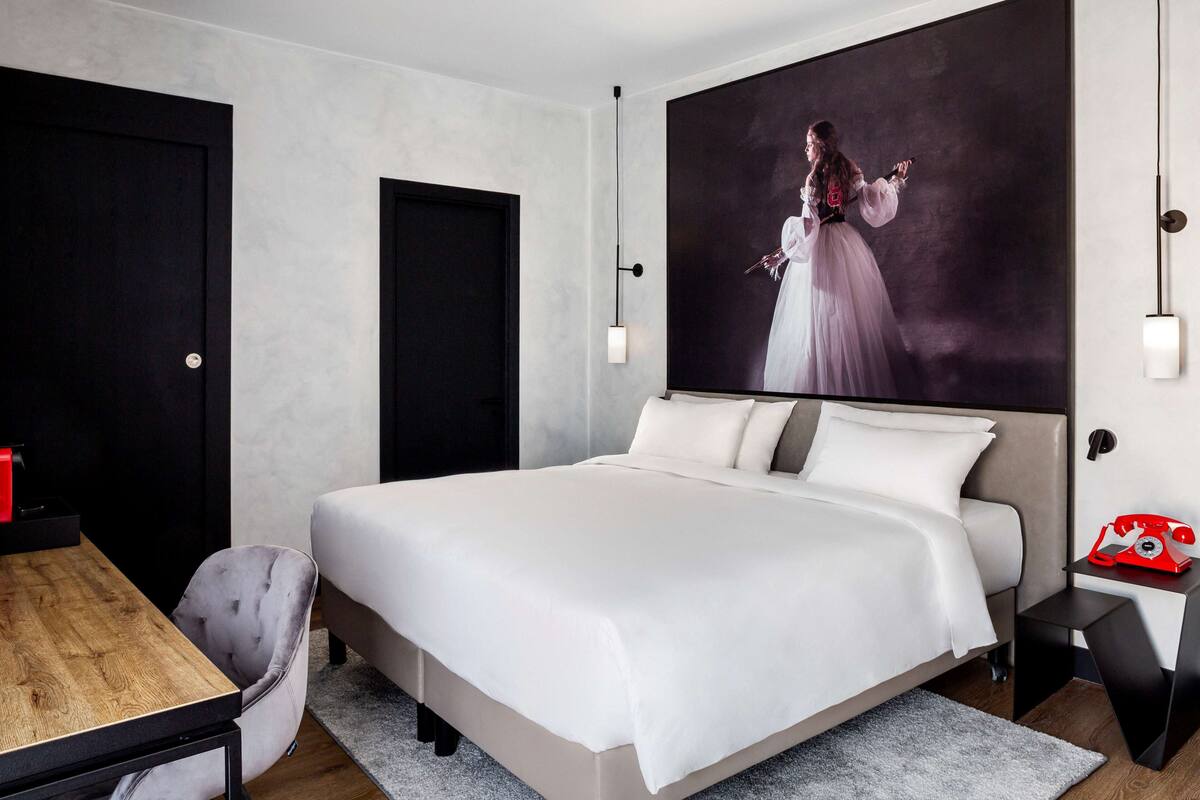 Een moderne hotelkamer met een groot bed, een houten bureau, een stoel, een nachtkastje met een rode telefoon en een groot schilderij aan de muur.