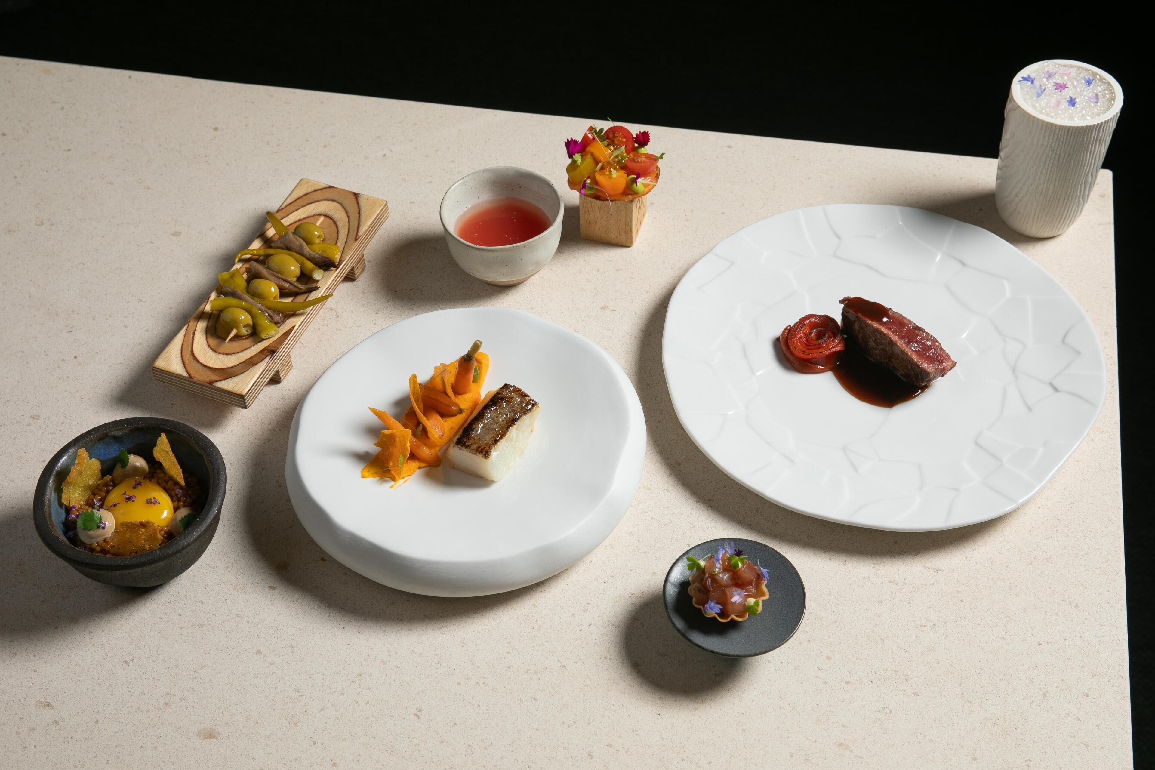 A table with various dishes, including a bowl of soup, a plate with a piece of meat, and a small dish with vegetables.