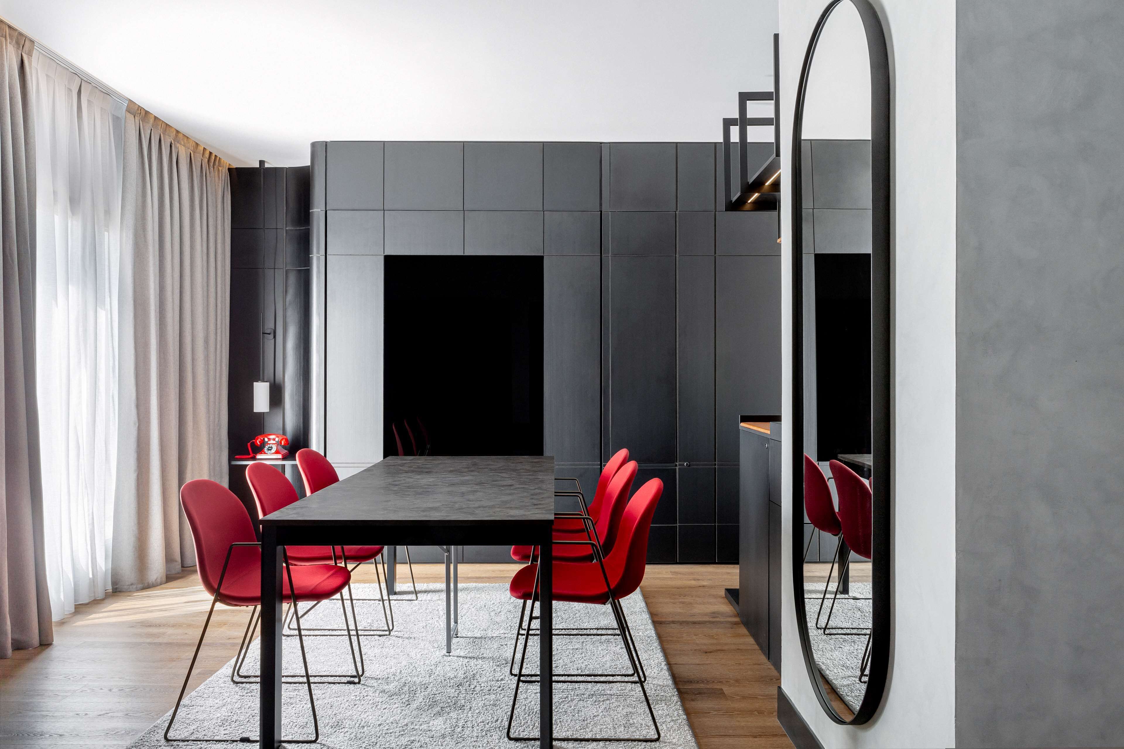 A modern dining area with a rectangular table, red chairs, a grey rug, and a large mirror.
