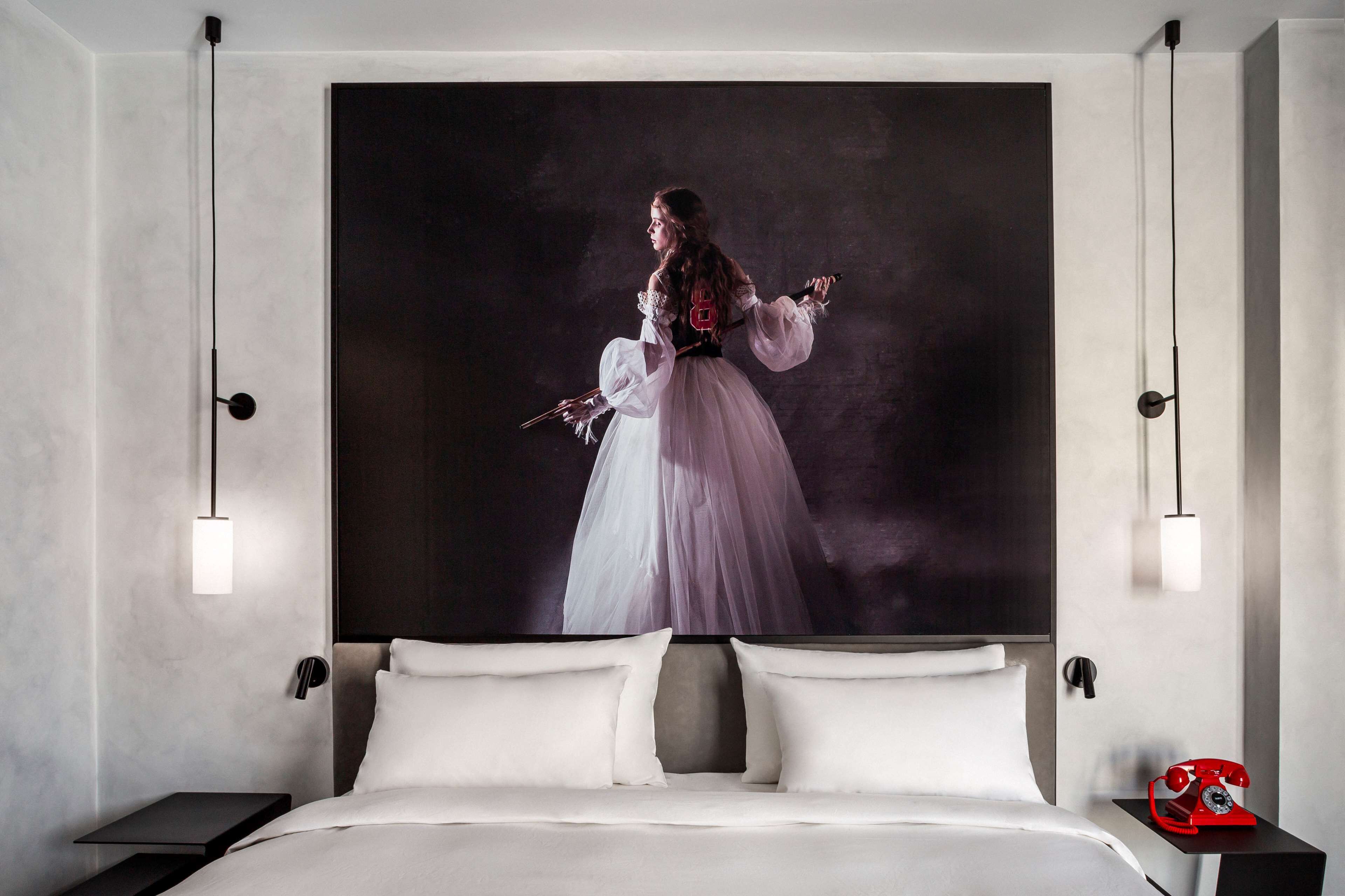 Une chambre d’hôtel moderne avec un grand lit, une table de chevet, un téléphone à cadran rouge et un grand portrait encadré accroché au mur.