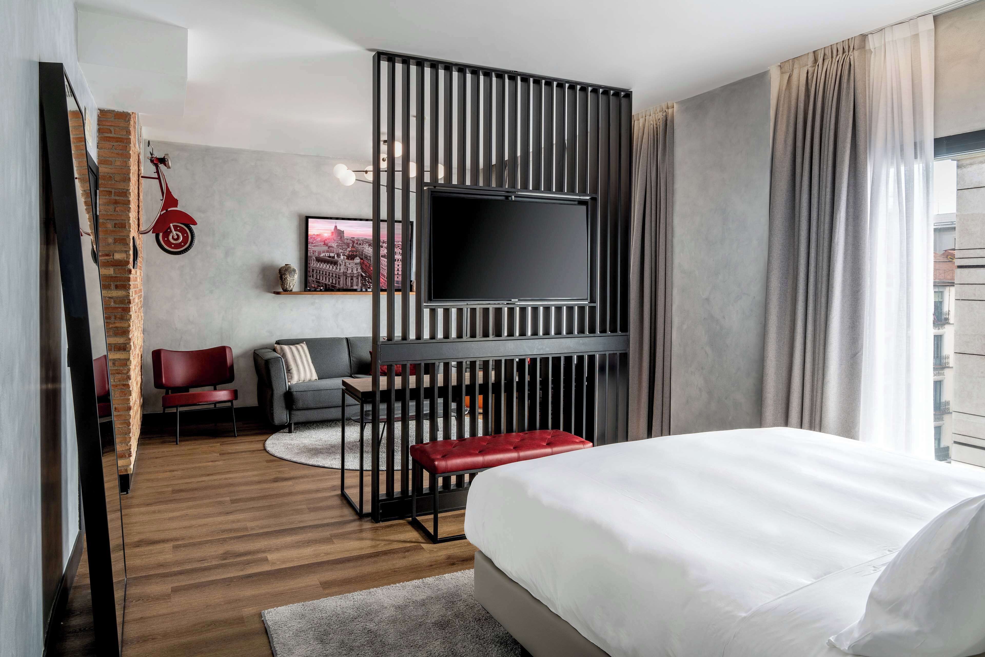 A modern hotel room with a large bed, a TV mounted on a wall, a red scooter decoration, and a cityscape painting.