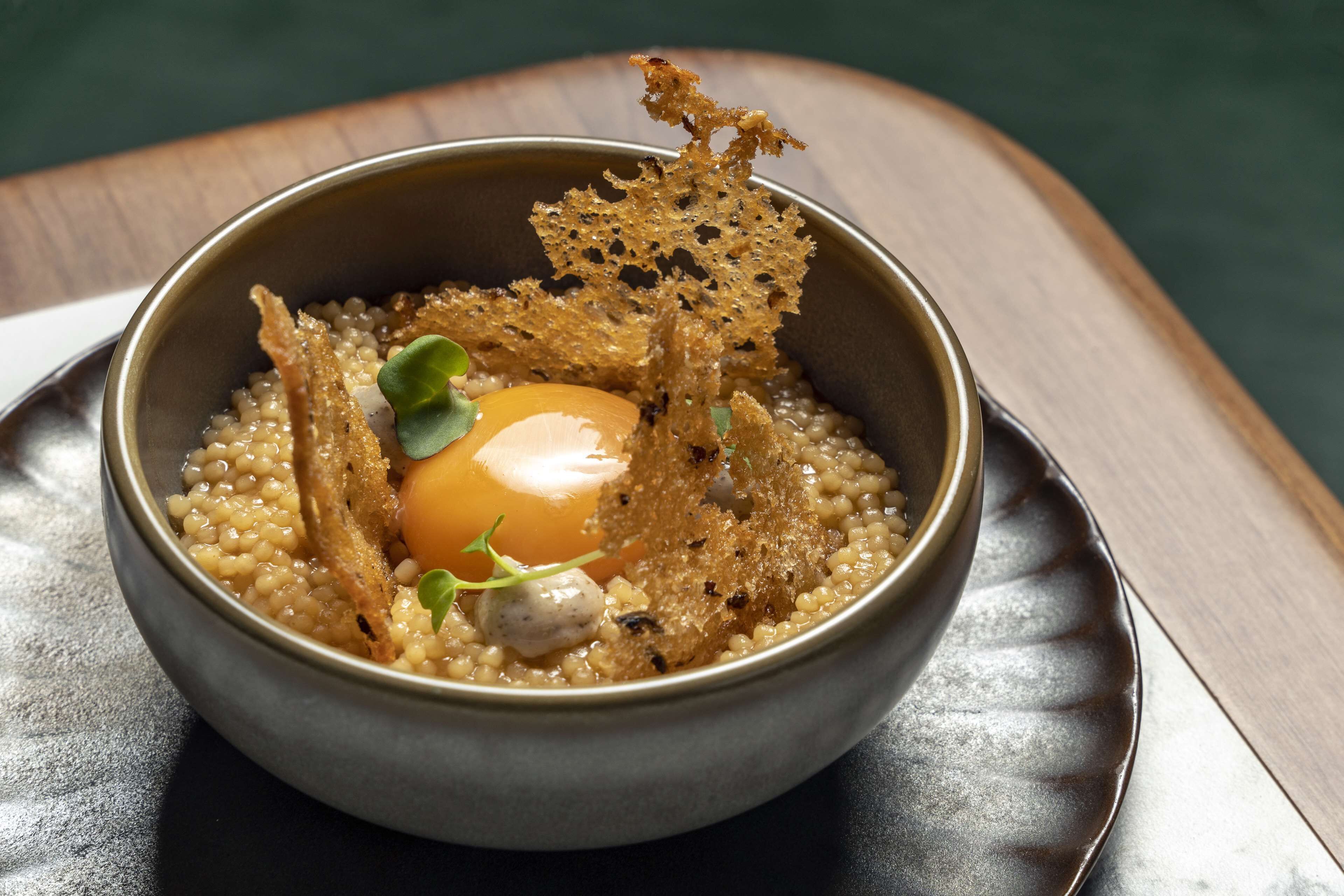 A bowl of food with a poached egg, grains, and crispy elements on a dark plate.
