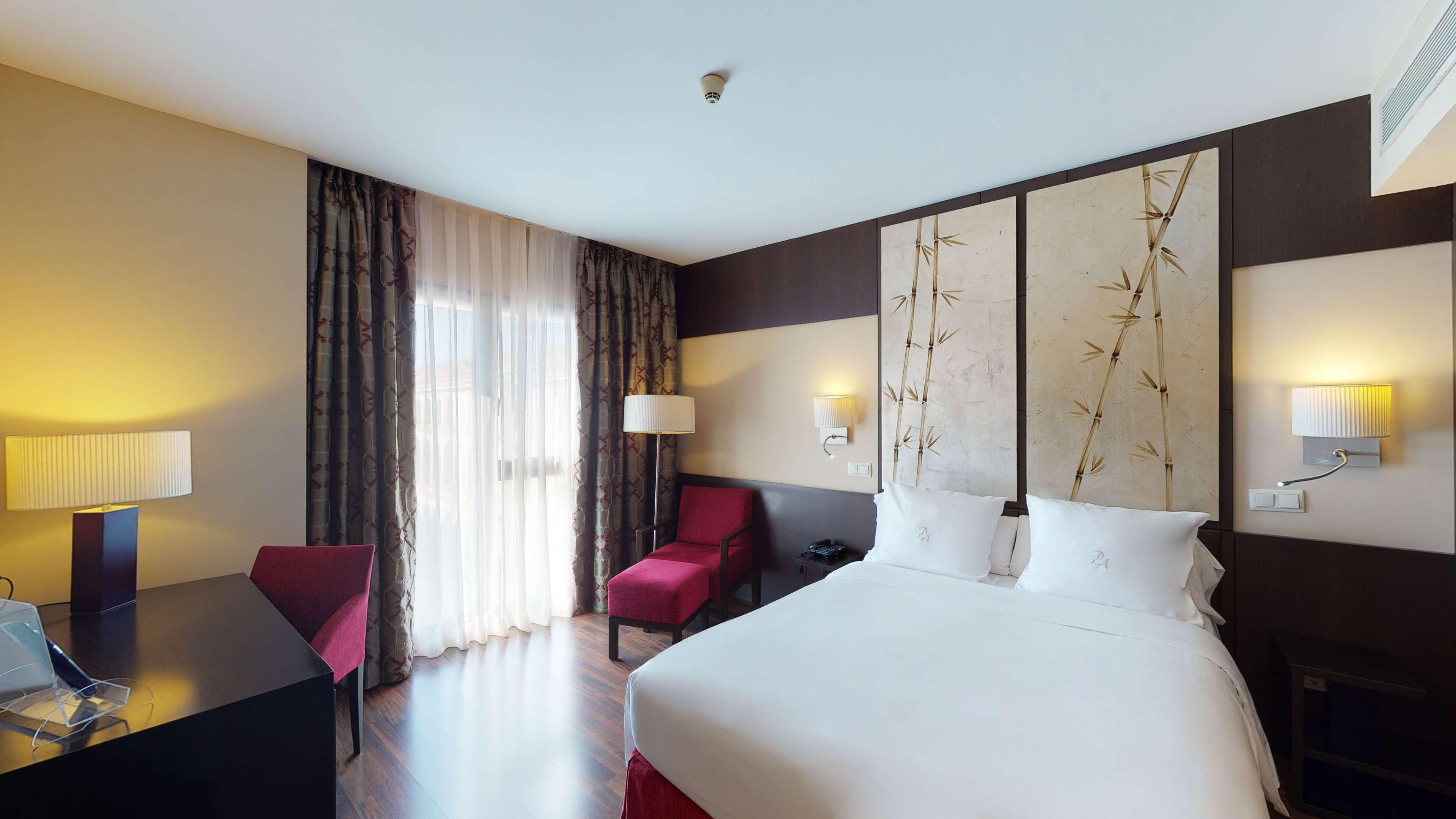 A hotel room with a bed, a desk, a chair, a bedside table, a lamp, a wall-mounted light, and a window with curtains.