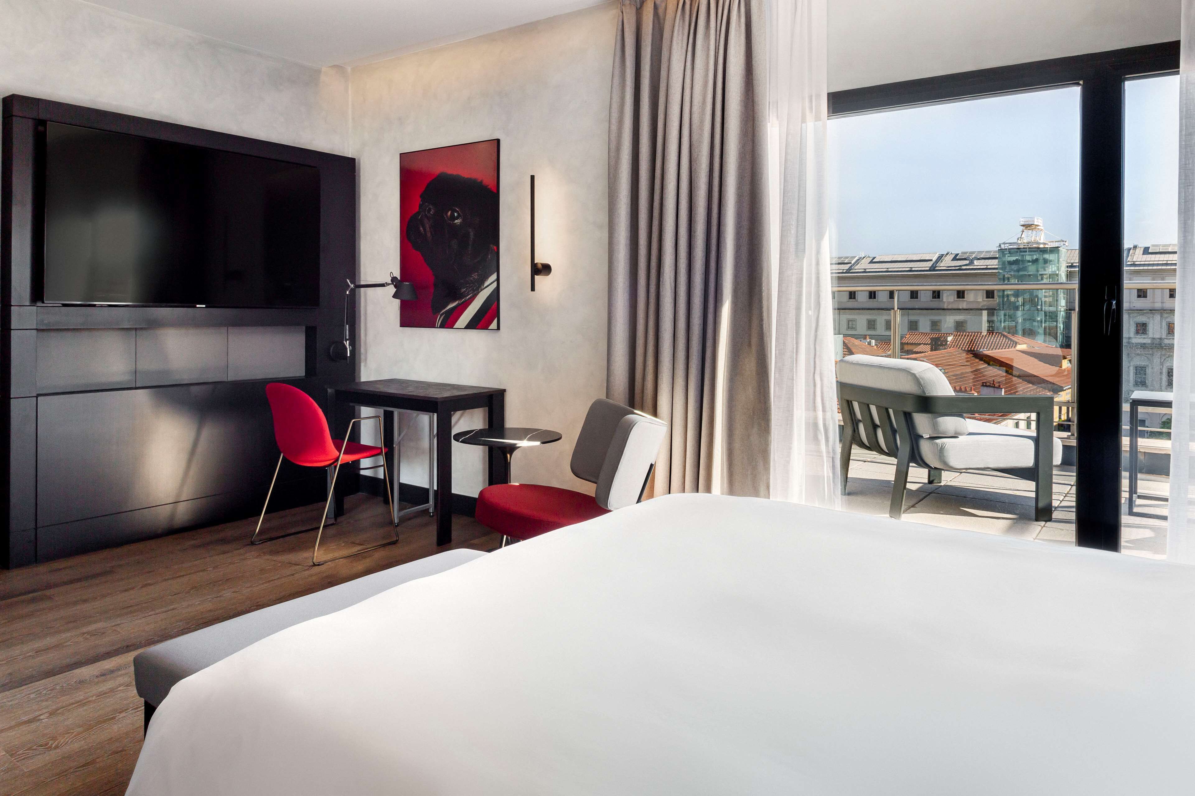 Una moderna camera d'albergo con un letto grande, una TV a schermo piatto montata a parete, un tavolino con due sedie e un'ampia finestra con vista sulla città.