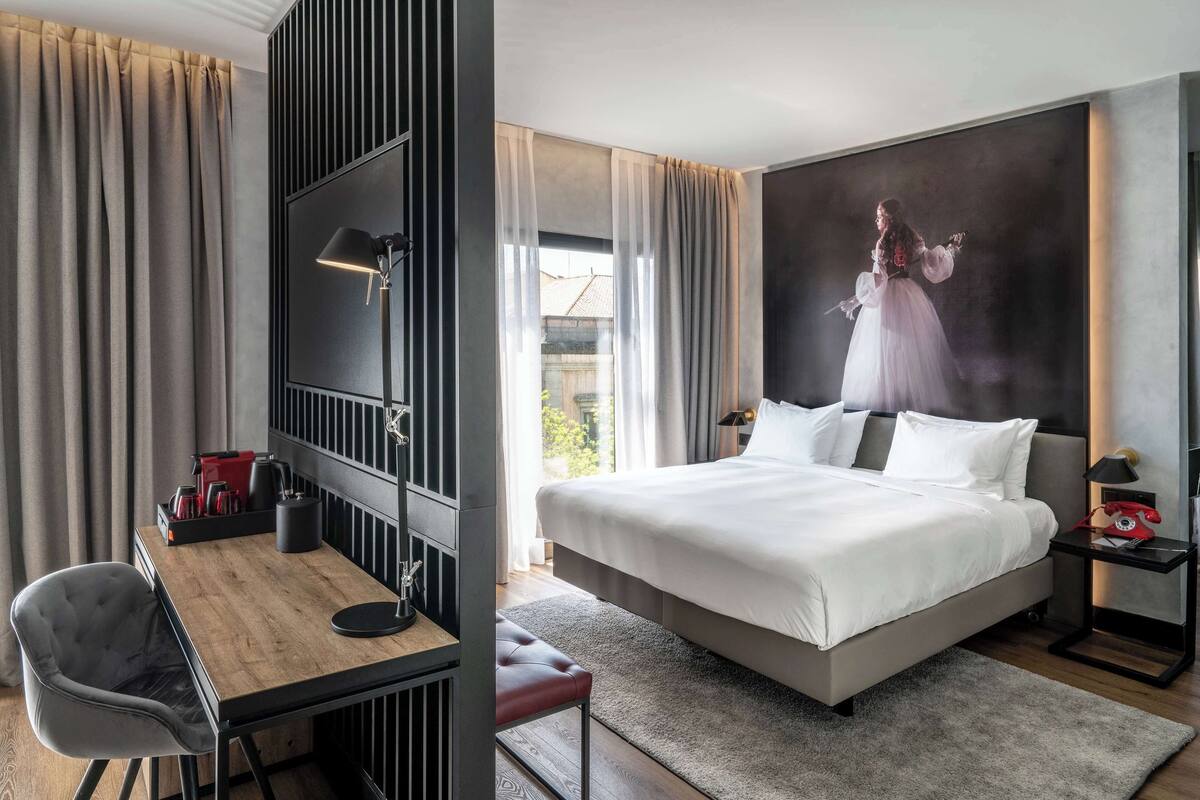 Een moderne hotelkamer met een groot bed, een bureau en een stoel. Er is een televisie aan de muur bevestigd, een rode telefoon en een klein tafeltje met een lamp.
