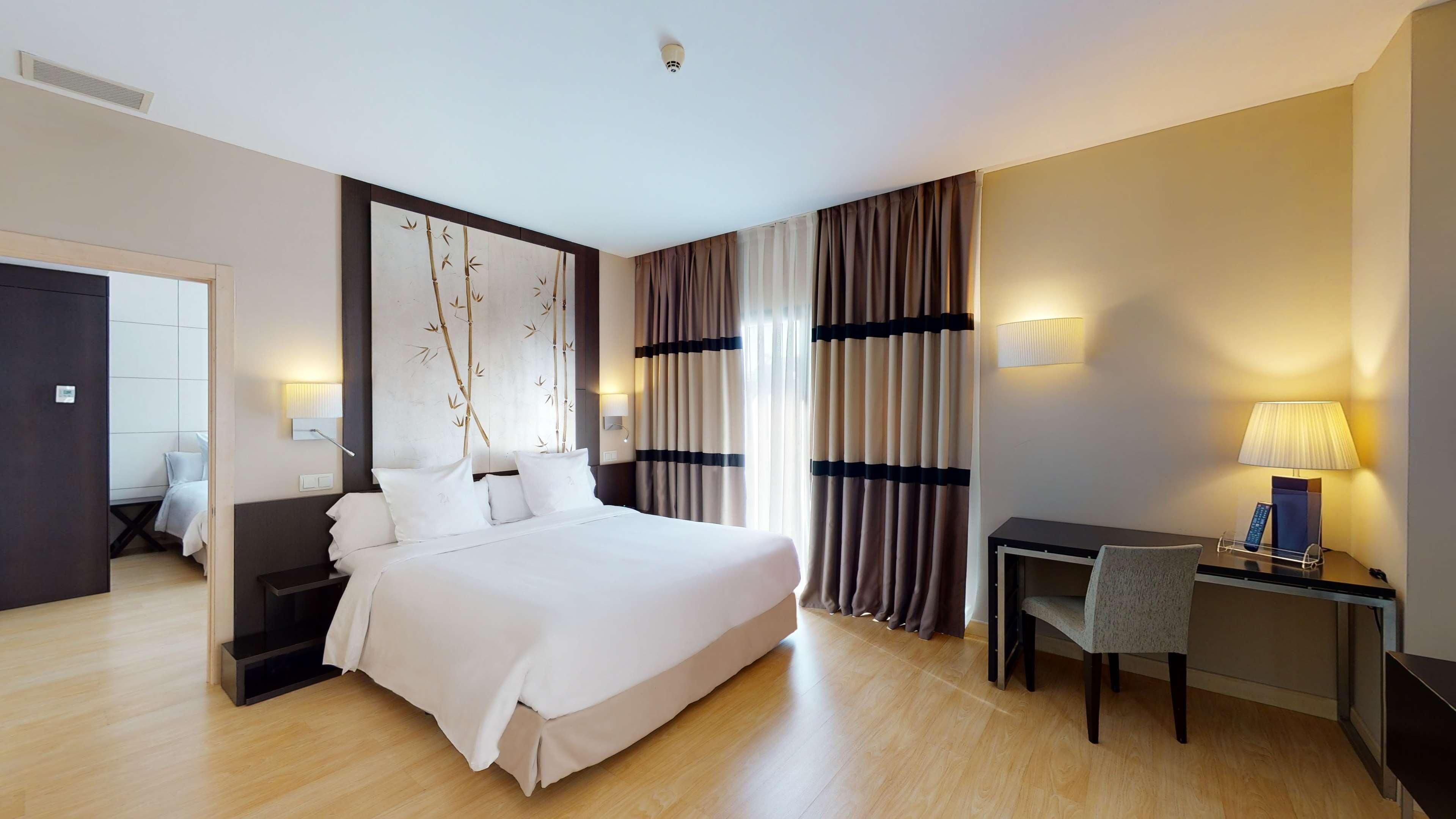 A hotel room with a bed, bedside tables, a desk, a chair, and curtains.