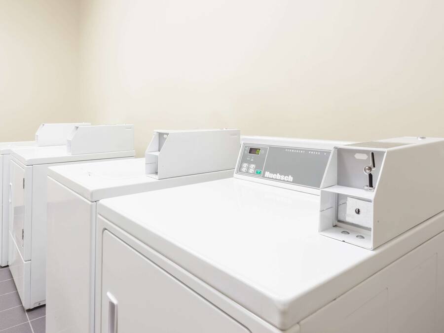A row of white, modern, front-loading washing machines in a laundry room with a plain wall and tiled floor.
