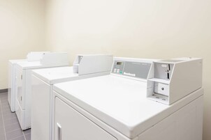 A row of white, modern, front-loading washing machines in a laundry room with a plain wall and tiled floor.