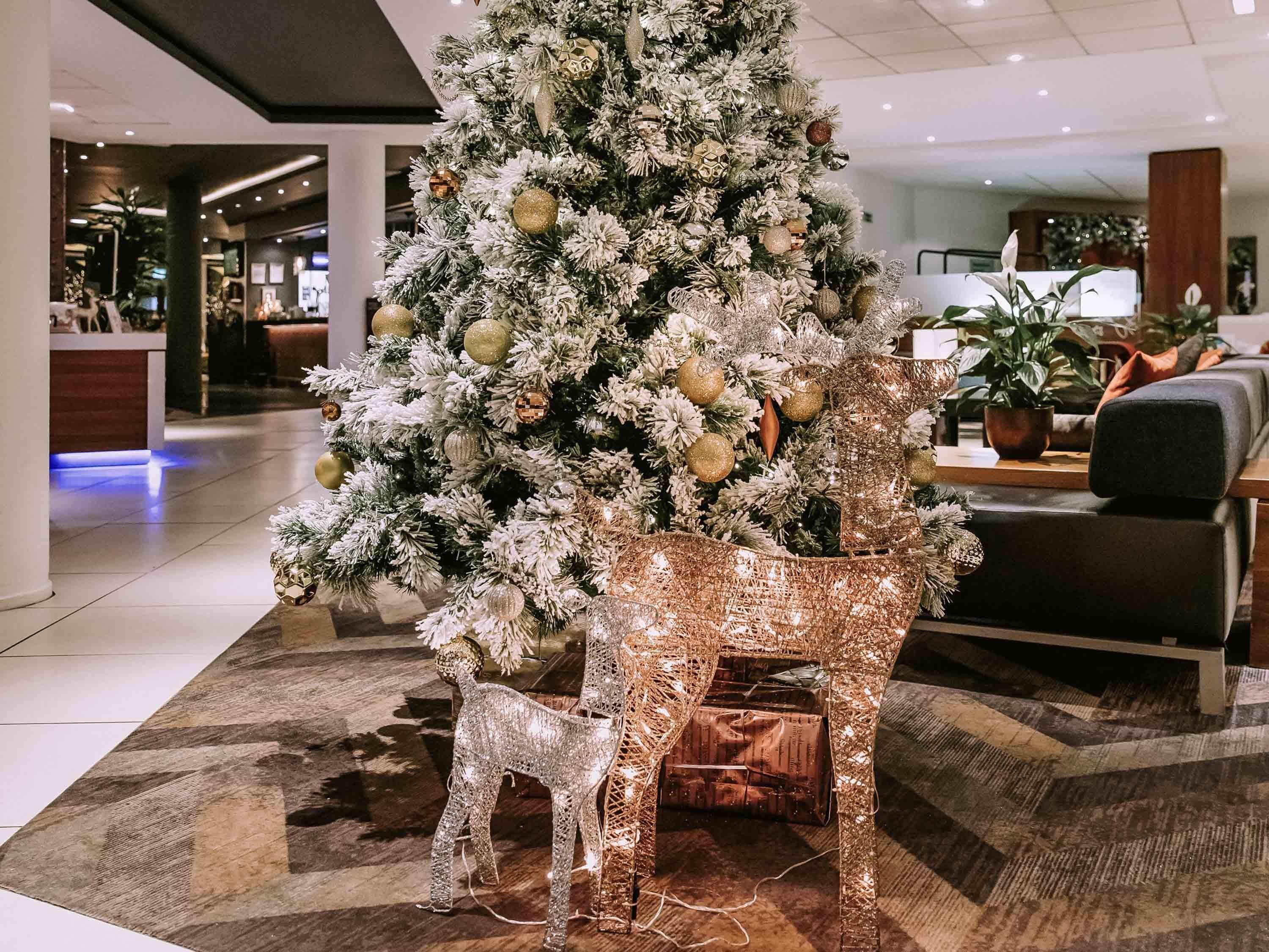 A Christmas tree with ornaments and lights, placed in a lobby area with a patterned carpet and seating.