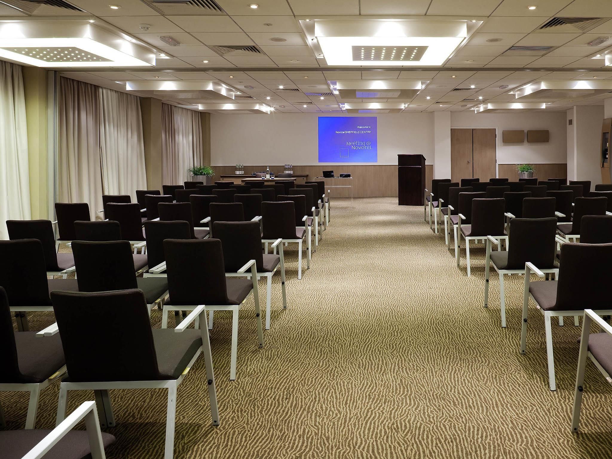 Un salón de conferencias con filas de sillas, un atril, una pantalla y suelo alfombrado.
